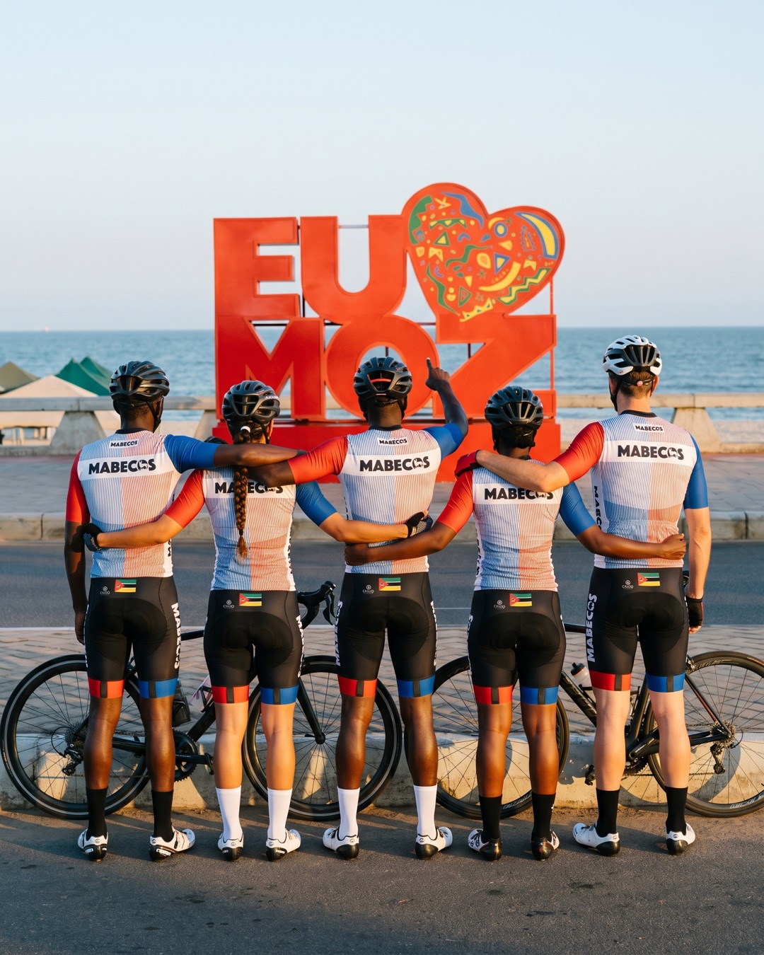 Cinco ciclistas Mabecos de costas, abraçados, em frente ao letreiro EU AMO MOZ na Marginal de Maputo
